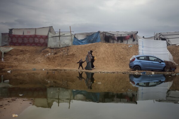 فلسطينيون نزحوا بسبب الهجوم البري الإسرائيلي على قطاع غزة يسيرون في مخيم مؤقت غمرته المياه في رفح، قطاع غزة، الأحد، 18 فبراير، 2024. (AP Photo/Mohammed Dahman)