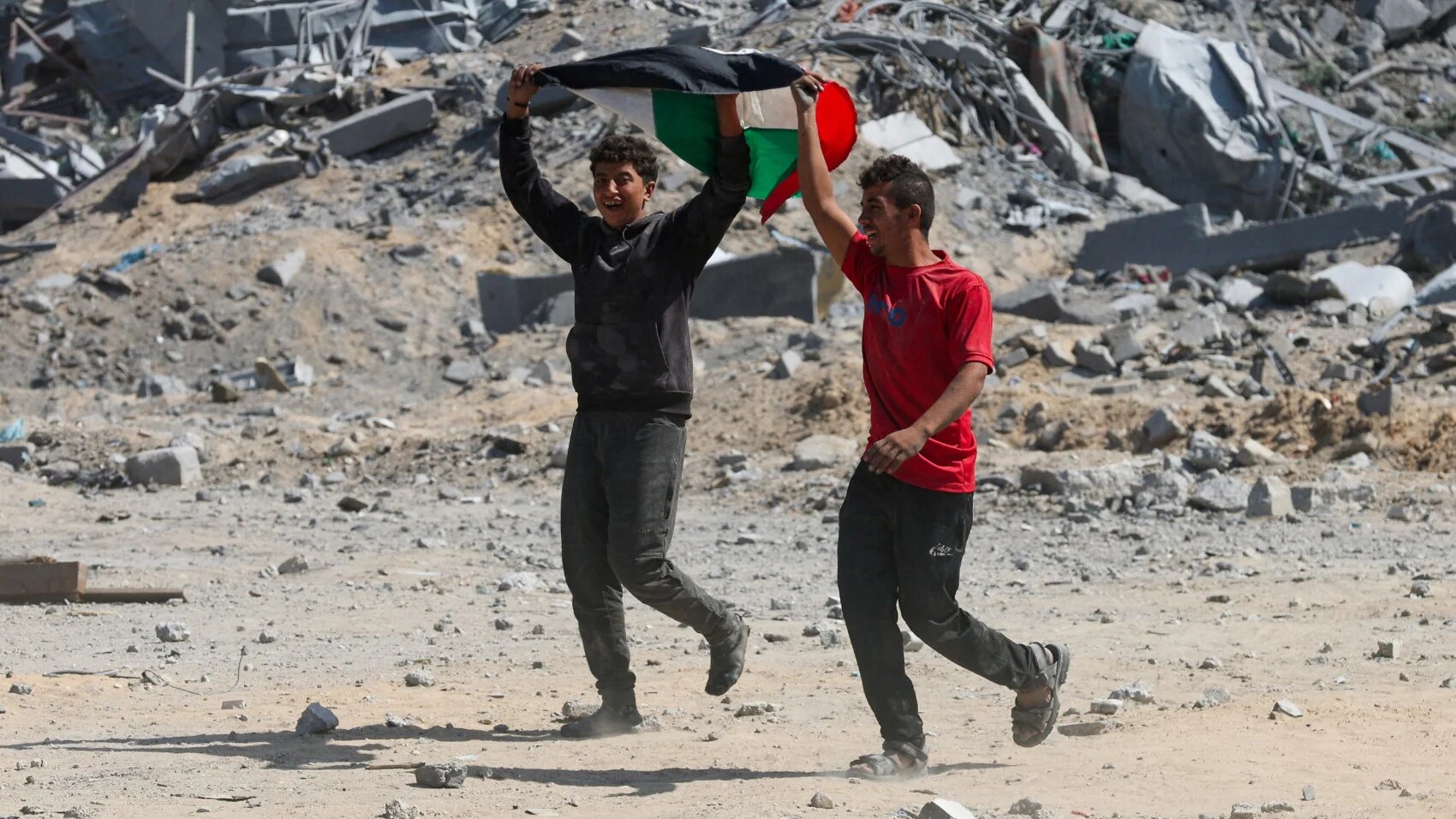 فلسطينيون يلوحون بعلمهم وهم يسيرون بين الأنقاض بعد انسحاب القوات الإسرائيلية من المنطقة، في خان يونس، جنوب قطاع غزة، 10 أكتوبر 2025 (رمضان عابد / رويترز)